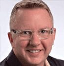 Portrait of a smiling man wearing glasses and a dark jacket.